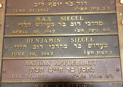 Image of a closeup on a Yahrzeit board, showing plaques commemorating David Leef (June 21, 1957), Max Siegel (April 18, 1947), Benjamin Siegel (June 20, 1947), Nathan Appleblatt (July 27, 1957) and Max Karlow (date cut off in image). Below each name is Hebrew lettering, a date in English, and more lettering to the right. The plaques are made of some kind of red-ish metal, and the lettering is golden. To the left, a semi-cut off light is visible next to David Leef's plaque.