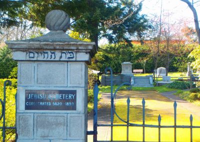 Cropped image of the entrance to the Jewish Cemetery of Victoria. A stone post and part of an old metal gate can be seen in the foreground. The post has a decorative, circular top. Below this, Hebrew writing is etched into the stone. Along the front of the post is a metal plaque that reads 'JEWISH CEMETERY / CONSECRATED 5620-1859'. The cemetery can be seen in the background, it is a sunny day and several grave markers stand in the background. A large tree looms to the left and shrubbery can be seen throughout the background.