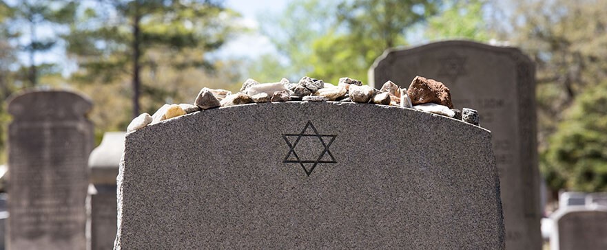 There is a domed-tablet headstone in a cemetery with a Star of David motif in the top-centre, but no visible names or other engravings, there are stones on top of the marker. The background contains blurred headstones and trees.