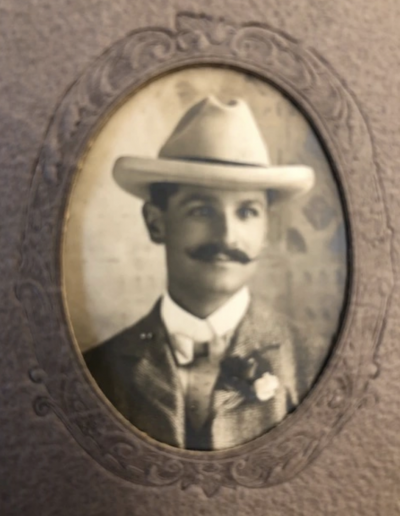 Image of a 19th century sepia-toned portrait within a brown oval frame. The man in the portrait sports a moustache, a 'Western' style hat, and a suit with a flower on its lapel. He looks off slightly to his left, with a gentle smirk on his face.