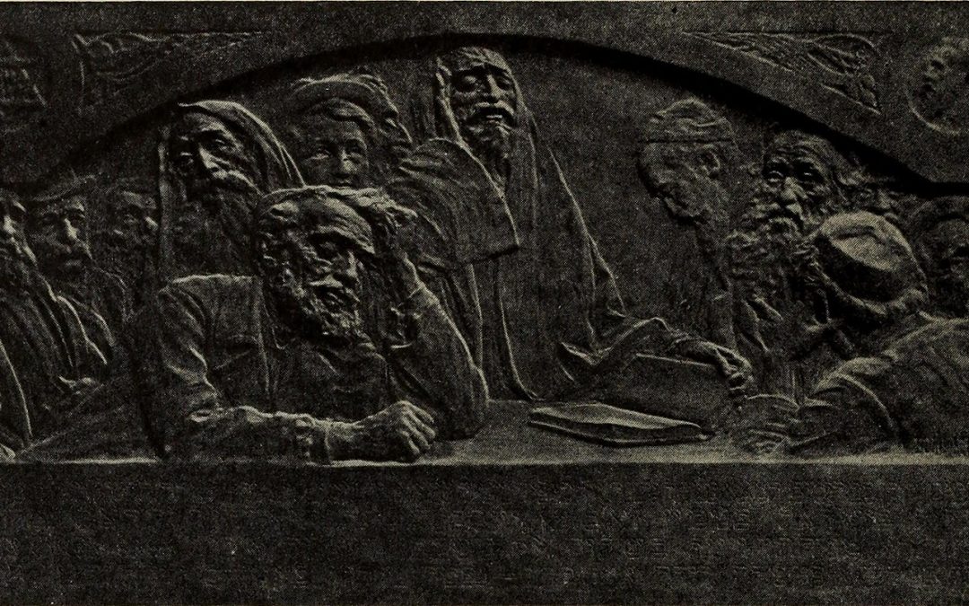 Image of a bas-relief in dark grey rock. The relief depicts 12 individuals, visibly only bearded men and boys, in mourning. Several wear robes, kippot, and fabric over their heads. One holds a book open. The man in the foreground leans over a table with his eyes closed and hand on his head. Around the frame of the relief are four carvings: from left to right, a Star of David in a circle, two unclear triangular designs, and a man's head in profile view (also in a circle).