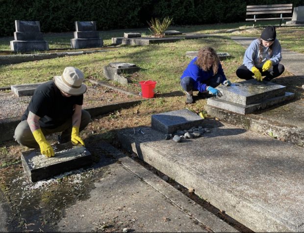 Three individuals squat beside gravestones. They are wearing gloves and using brushes to clean the granite graves.