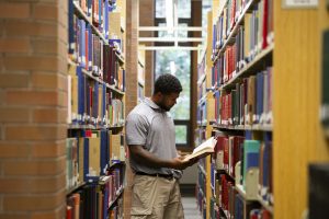 The image shows a student reading a book in one of the rows of the Diana M. Priestley Law Library at UVic Law.