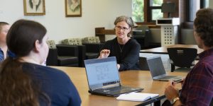 The photo shows a professor addressing students sitting around a table in the library.