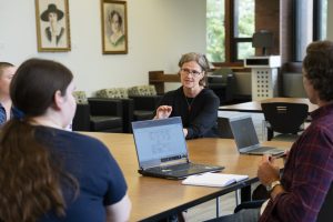 The photo shows a professor addressing students sitting around a table in the library.