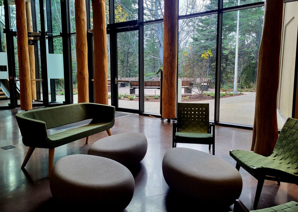 The photo shows an indoor seating area in the new Indigenous Law wing with green seating and grey ottomans. In the distance, a wooded area and outdoor seating can be seen.