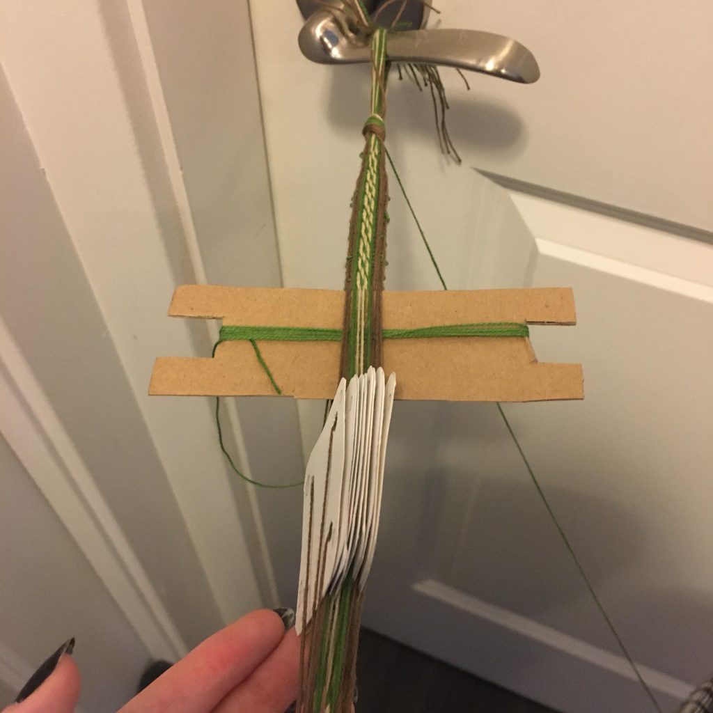 This image shows the end of the weave tied to a silver door handle, the cardboard shuttle between the threads to hold them still for a picture, the tablets arranged vertically, and a hand with black nails holding it up.