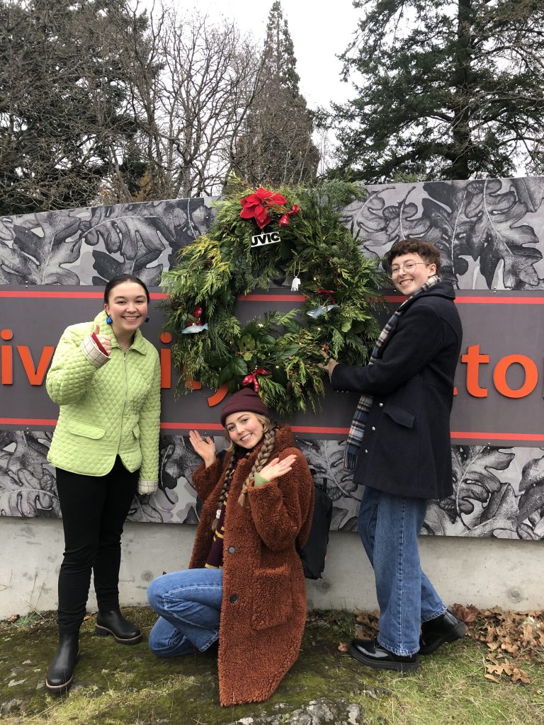 Three Uvic students posing beside a DIY wreathe that is hanging from the Uvic sign. 