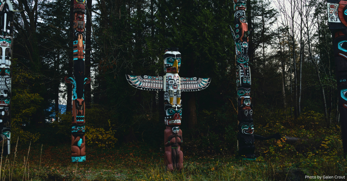 Three intricately carved and painted totem poles standing in a tranquil forest, their vibrant colors of red, black, white, and turquoise contrasting with the muted tones of the surrounding trees and foliage. The central totem pole features an extended wing-like structure. The ground is covered with grass and fallen leaves, indicating a fall season.