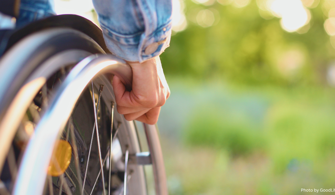 Close-up of a person’s hand gripping the wheel of a wheelchair, wearing a denim jacket, with a blurred greenery background.