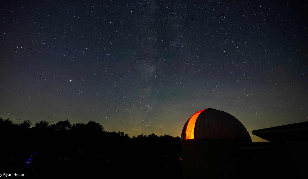 A serene night scene featuring an observatory with a glowing red dome, set against a backdrop of a starlit sky with the Milky Way galaxy distinctly visible, and silhouetted trees in the foreground. Photo credit is given to Ryan Heuer.