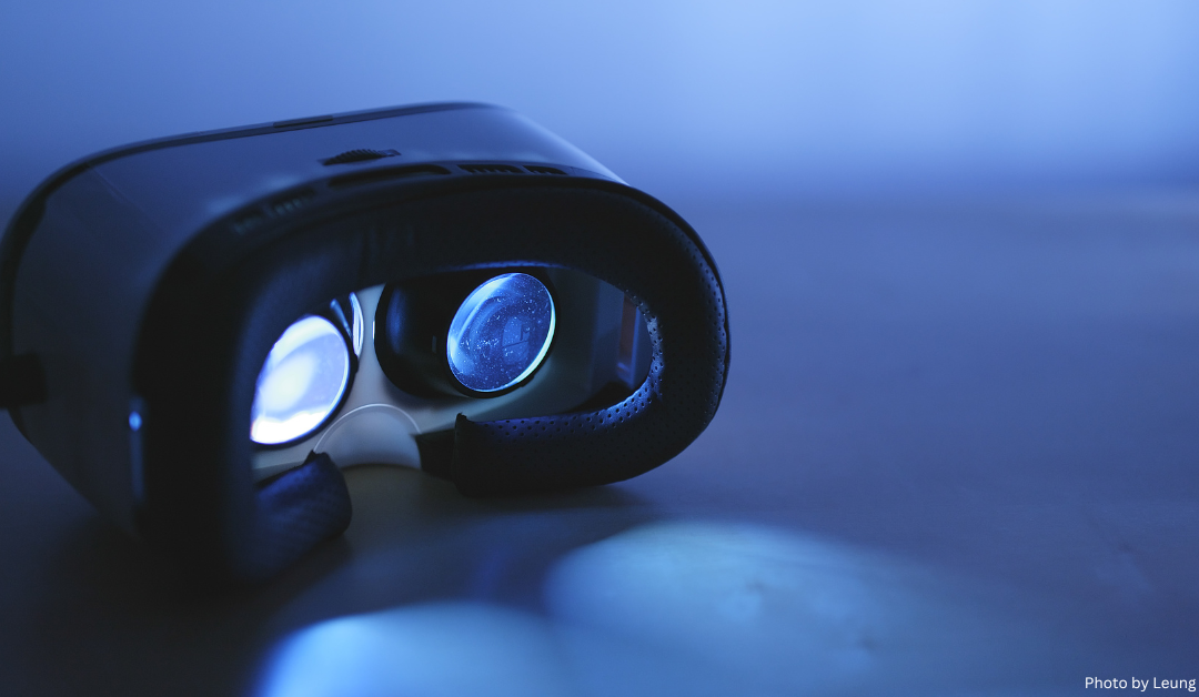 A close-up of a virtual reality headset against a blue background, with the front facing towards the right. The lenses of the headset are visible and reflect a white light, giving the image a futuristic feel.