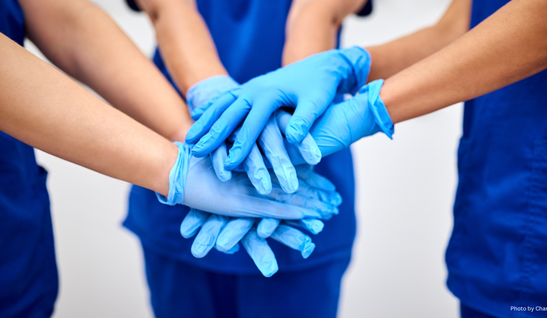 A group of nurses in blue scrubs stacking their gloved hands together as a symbol of teamwork.