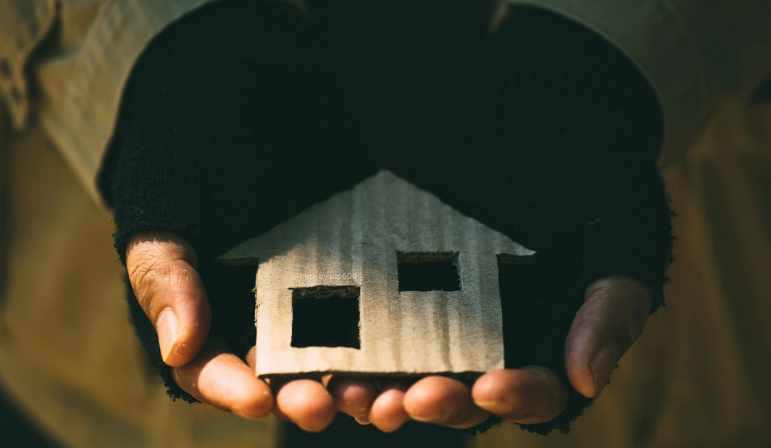 Hands wearing fingerless gloves holding a small, cardboard cutout of a house with two square windows, symbolizing themes of shelter or home. The image is dimly lit, giving it a warm, rustic, and contemplative feel.