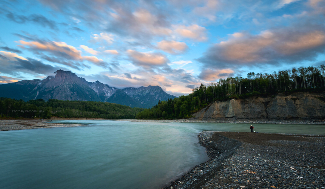 A serene landscape featuring the Skeena River flowing through a valley with a backdrop of majestic mountains under a partly cloudy sky. The riverbank is lined with trees, and a person is standing near the water's edge on the right side of the image. The sky is painted with soft hues of blue and pink from the setting or rising sun.