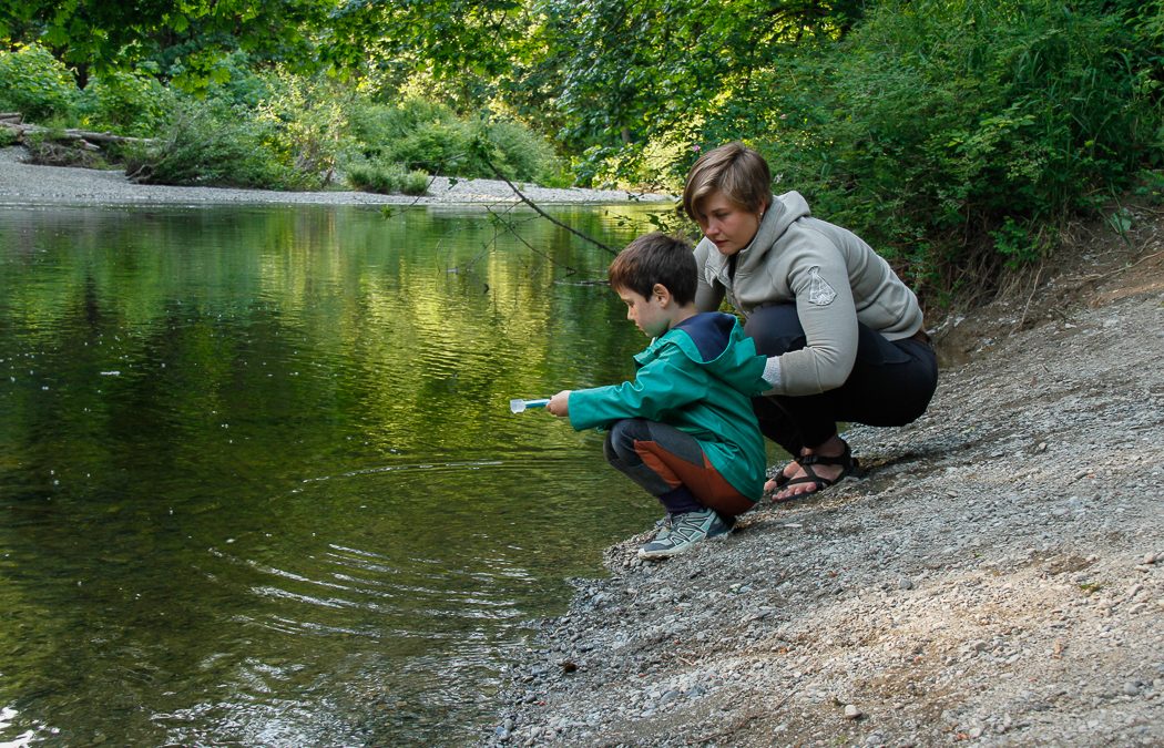 Kristina watches a child use a water monitoring tool near a still river landscape